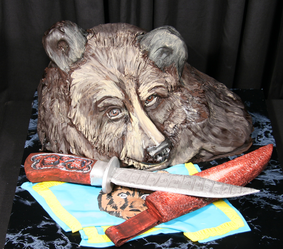 A realistic cake sculpted to resemble a brown bear's head rests behind a detailed cake replica of a knife with a red handle and sheath, all displayed on a black marble-patterned surface.