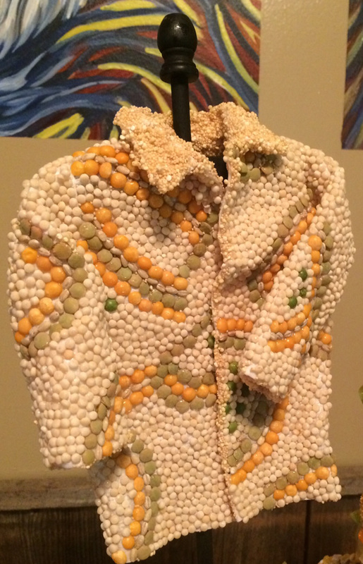 A miniature jacket sculpture crafted from white, orange, and green sugar beads is displayed on a black stand against a backdrop of abstract art.