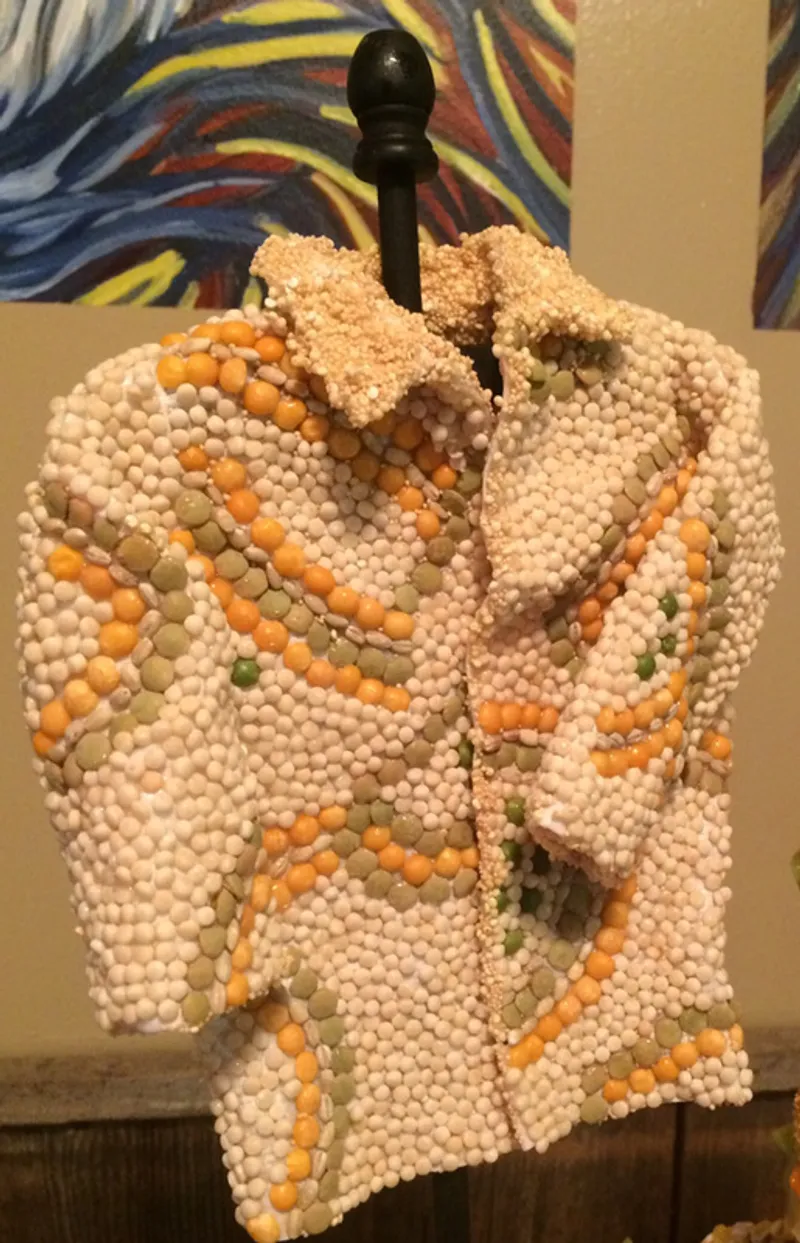 A miniature jacket sculpture crafted from white, orange, and green sugar beads is displayed on a black stand against a backdrop of abstract art.