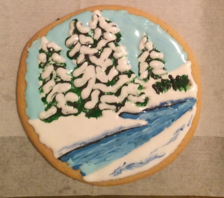 A round sugar cookie decorated with royal icing to depict a winter landscape featuring snow-covered pine trees, a blue sky, and a winding blue river.
