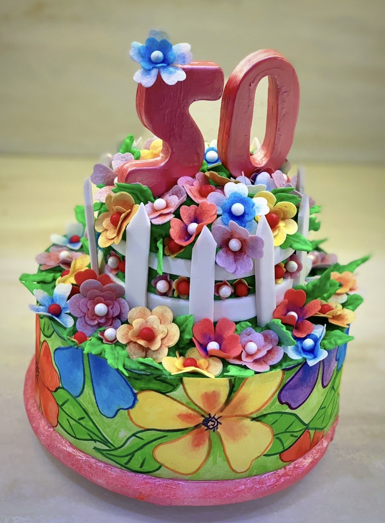 A 50th birthday cake with folk-art-inspired style featuring a large glossy pink "50" topper crowned by a delicate blue-and-white sugar flower, two tiers — lower tier with painted green base and bold stylized flowers in yellow, orange, blue, purple, upper tier encircled by miniature white picket fence with dimensional fondant flowers in coral, lavender, mustard, sky blue.