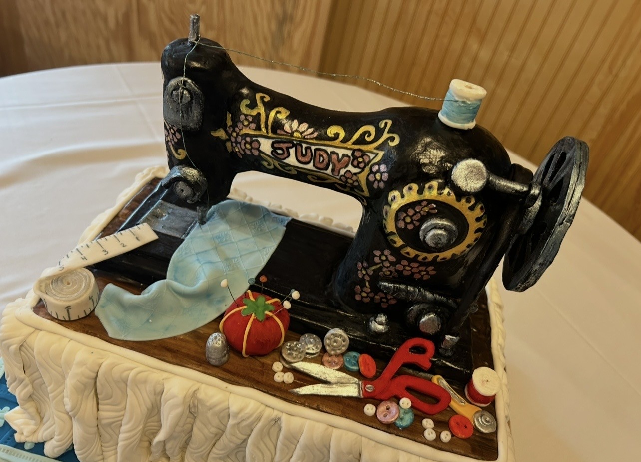 A sculpted cake designed as a vintage black sewing machine with glossy finish and intricate gold and floral detailing, the name "JUDY" scripted in white, spool of light blue thread with trailing thread, surrounded by miniature fondant sewing accessories including red pincushion with pins, red-handled scissors, buttons, thimble, measuring tape, quilted fabric, on a base adorned with ruffled white fondant.