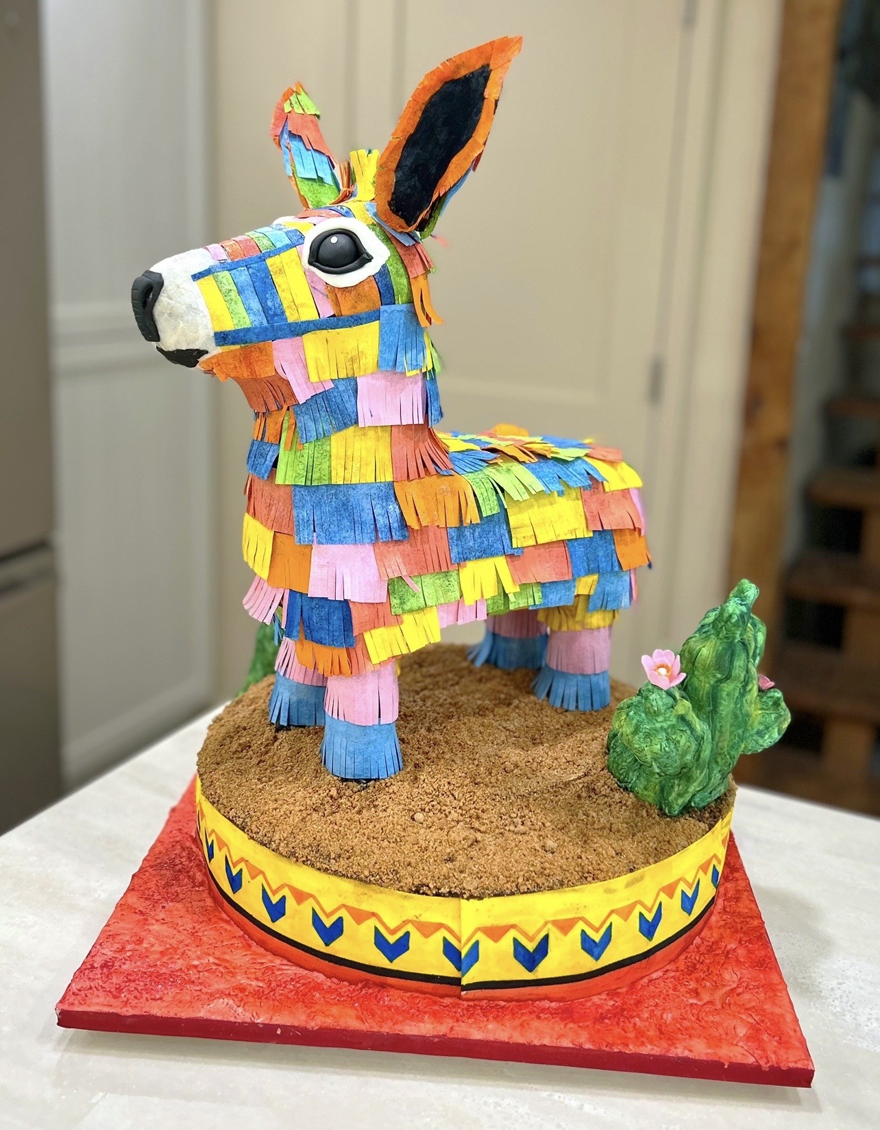 A piñata donkey cake adorned with colorful paper strips in shades of blue, yellow, pink, orange, and green, featuring a large expressive eye and detailed snout, sitting on a circular base covered in crushed graham crackers mimicking sand, surrounded by a geometric-patterned border and a small fondant cactus with a pink flower.