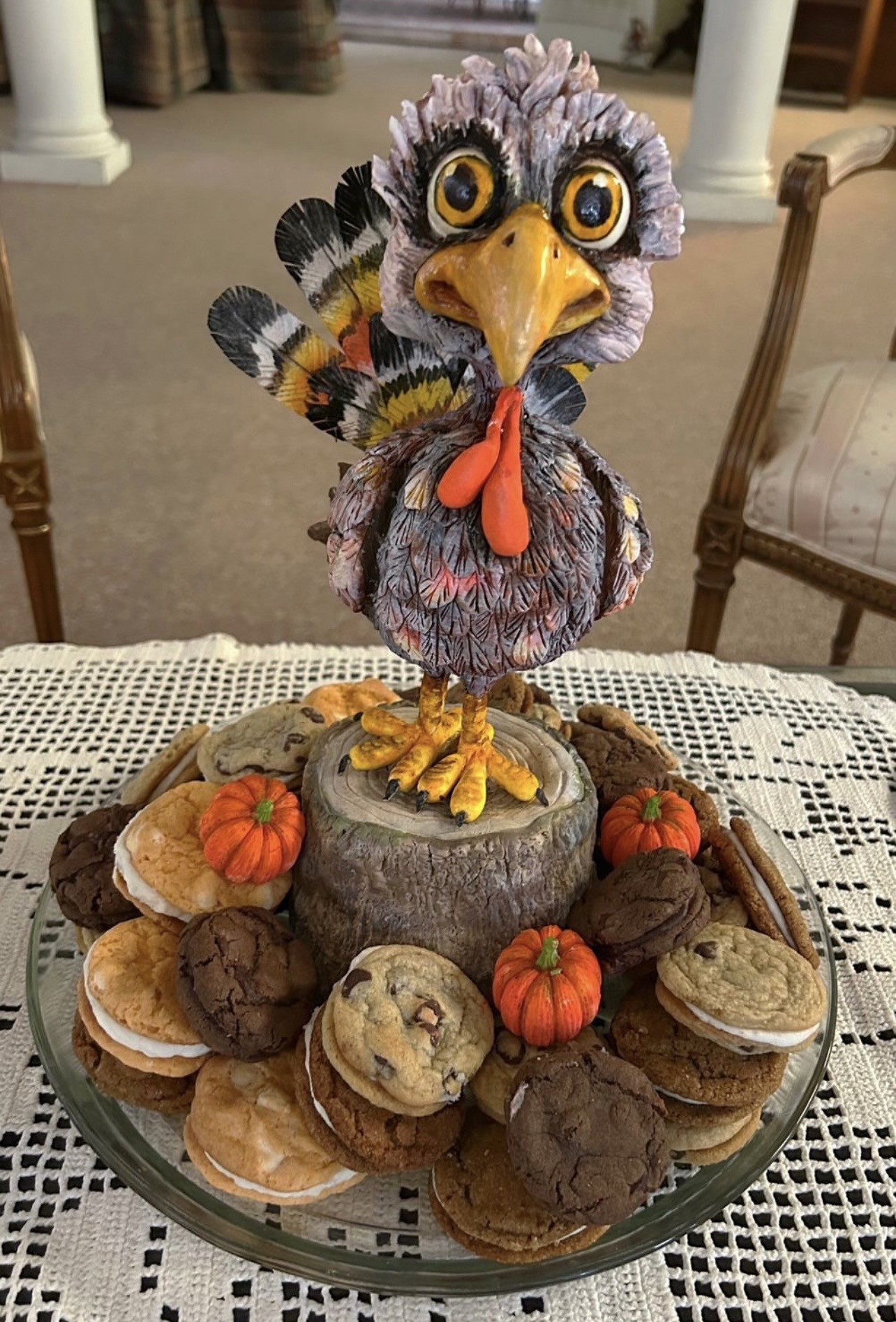 A Thanksgiving-themed cake featuring a large stylized turkey figurine with wide yellow eyes, bright orange beak and wattle, intricately detailed feathers in gray, brown, red, and orange fanning out as tail feathers, perched atop a faux tree stump surrounded by chocolate chip cookies, sugar cookies with white frosting, and small glossy orange pumpkins.
