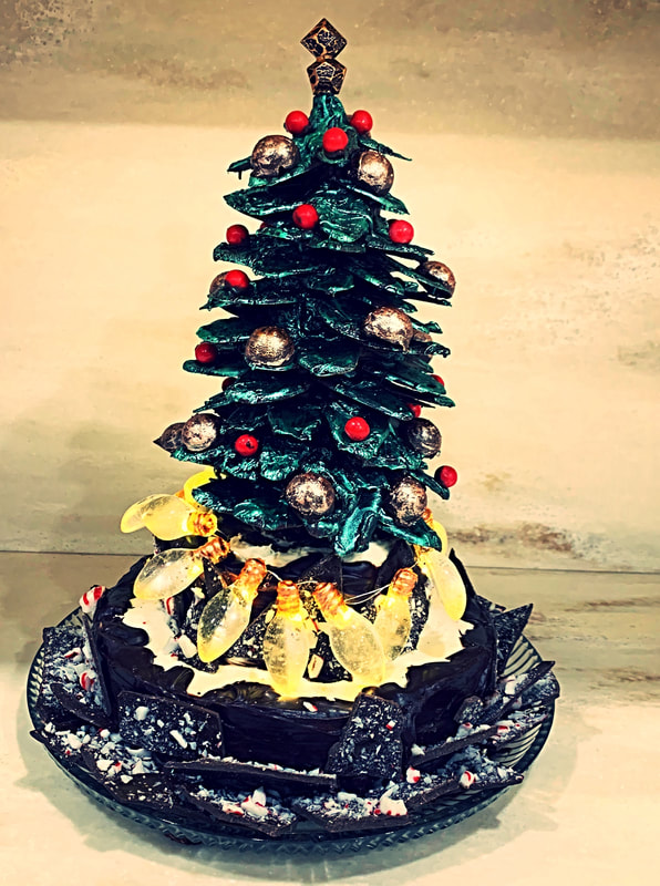 An elaborate Christmas tree cake features dark green piped frosting branches adorned with red and gold baubles, surrounded at the base by glowing yellow candy lights and resting on a chocolate-dusted plate.