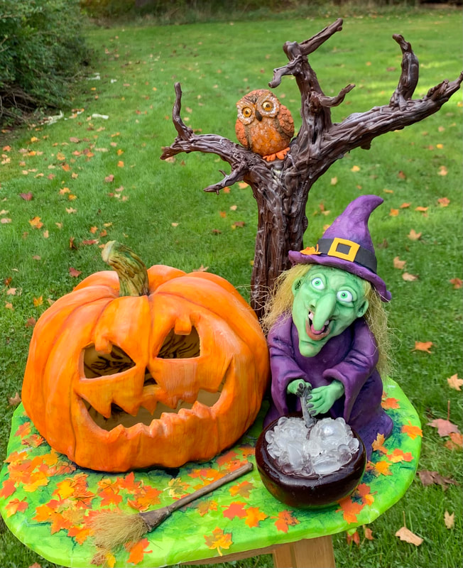 A Halloween display features a carved jack-o'-lantern, a green-skinned witch stirring a cauldron, and an owl perched on a tree branch, all set on a painted green base decorated with autumn leaves.