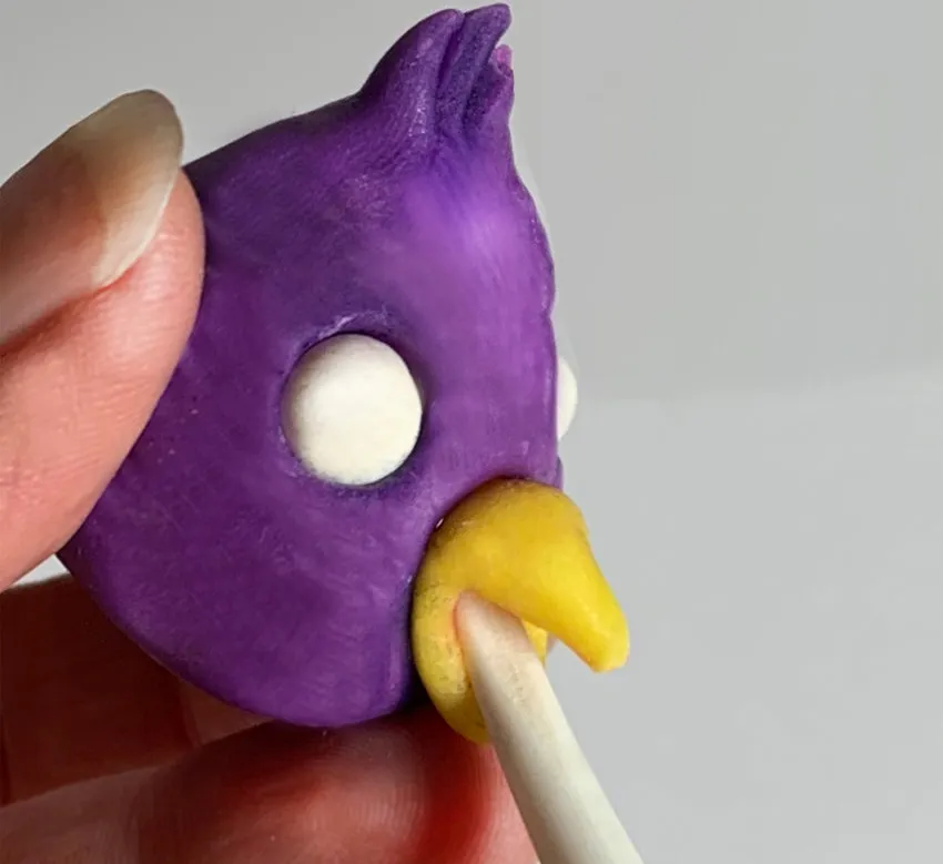 Hand shaping a yellow modeling chocolate beak with a clay tool.