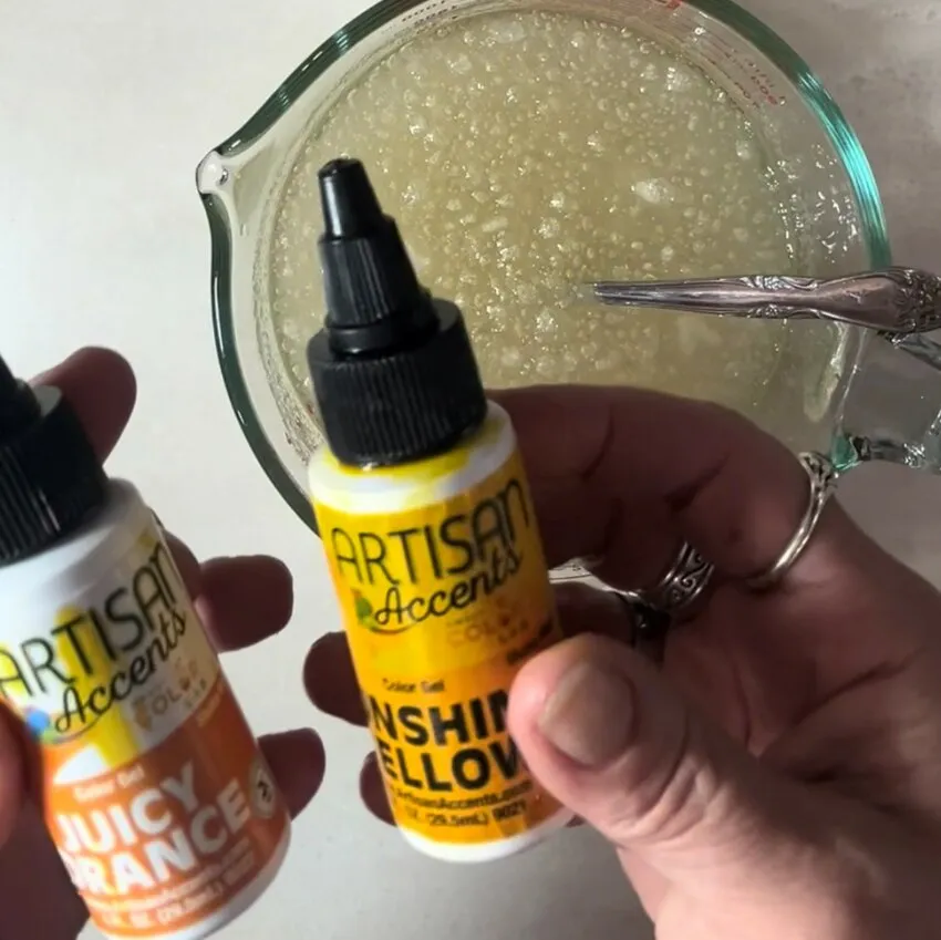 Hands hold orange and yellow colorant bottles beside the gelatin mixture.