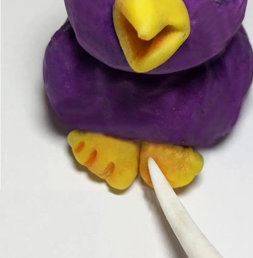 Hand detailing yellow modeling chocolate feet on the lovebird with a sculpting tool.
