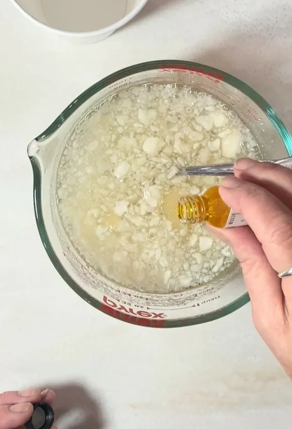 A hand pours candy flavoring oil into the gelatin mixture in a glass cup.