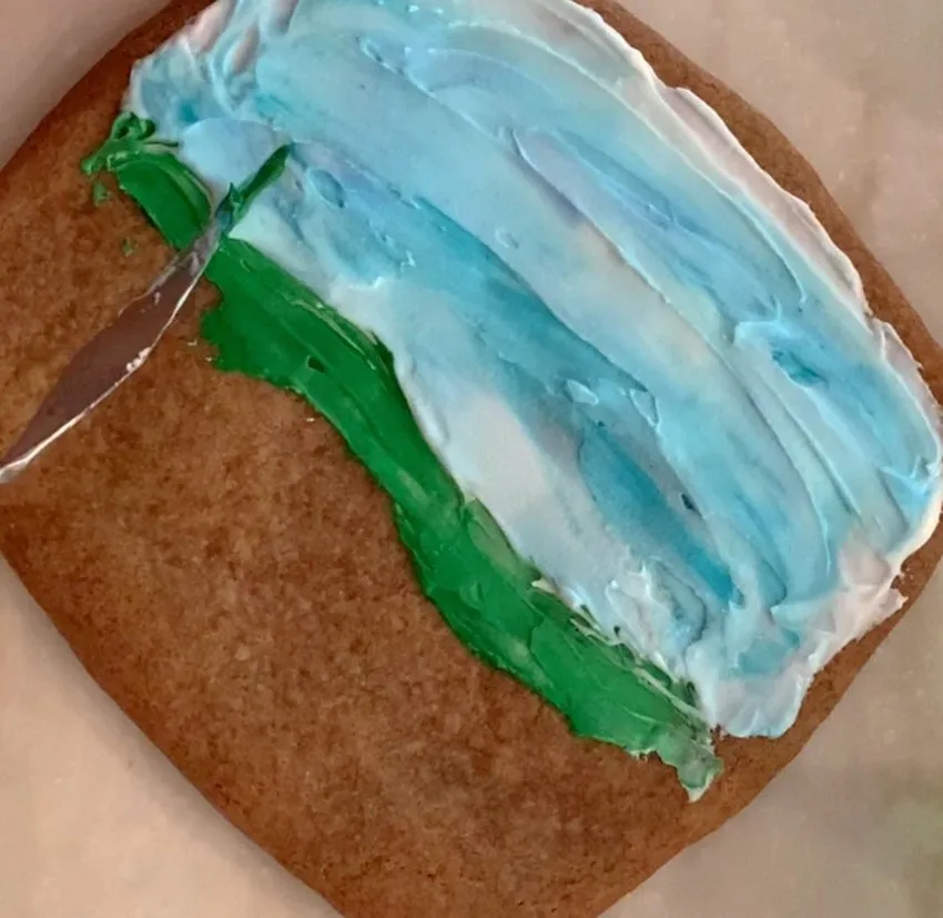Knife spreading green royal icing for grass on the cookie.