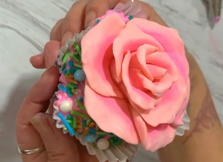 Hands holding a cupcake topped with a pink modeling chocolate rose and sprinkles.
