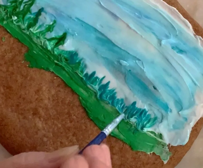 Hand using a brush to add green royal icing trees to the cookie.