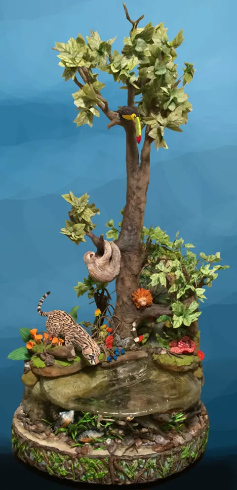A sculpted cake depicting an Amazon rainforest scene with a toucan, sloth, and jaguar around a stream.