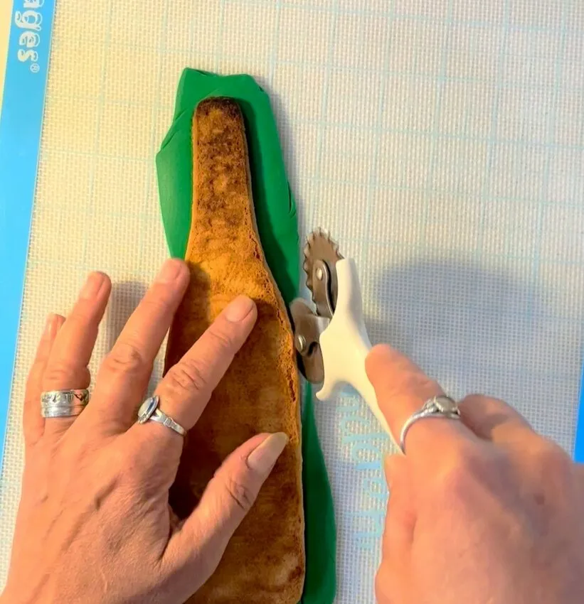 Hand trimming green modeling chocolate around a bottle-shaped cookie.