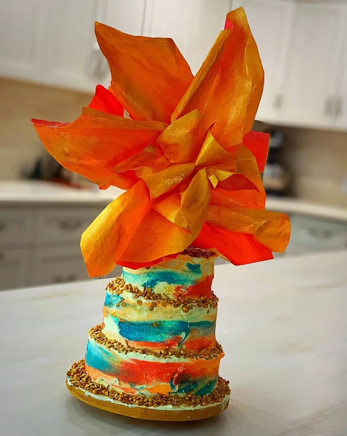 A three-tiered cake decorated with colorful watercolor-style frosting and gold sprinkles topped with an orange and red wafer paper flower