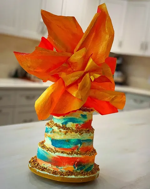 A three-tiered cake decorated with colorful watercolor-style frosting and gold sprinkles is topped with a large, vibrant orange and red flower made from edible wafer paper.