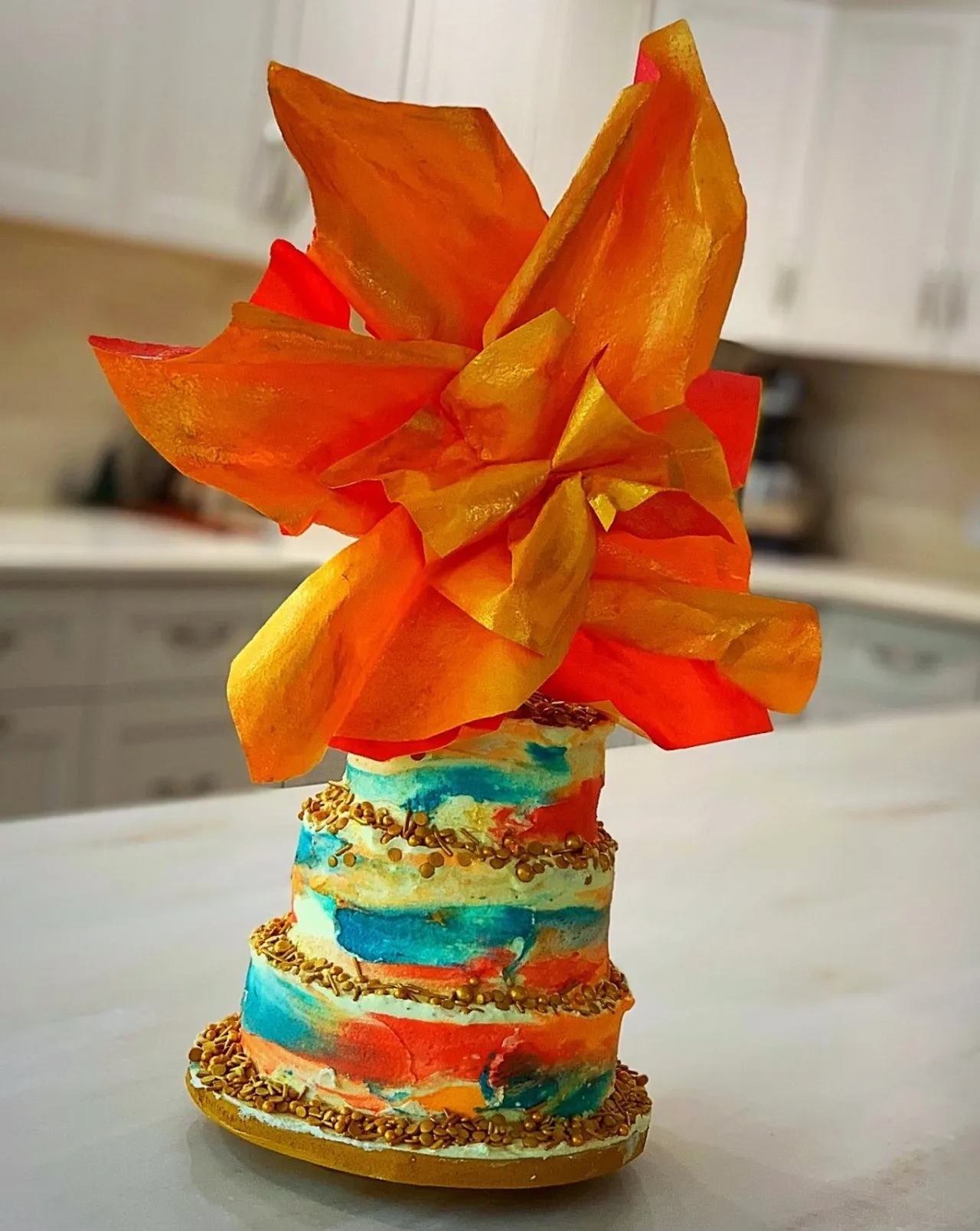 A three-tiered cake decorated with colorful watercolor-style frosting and gold sprinkles is topped with a large, vibrant orange and red flower made from edible wafer paper.