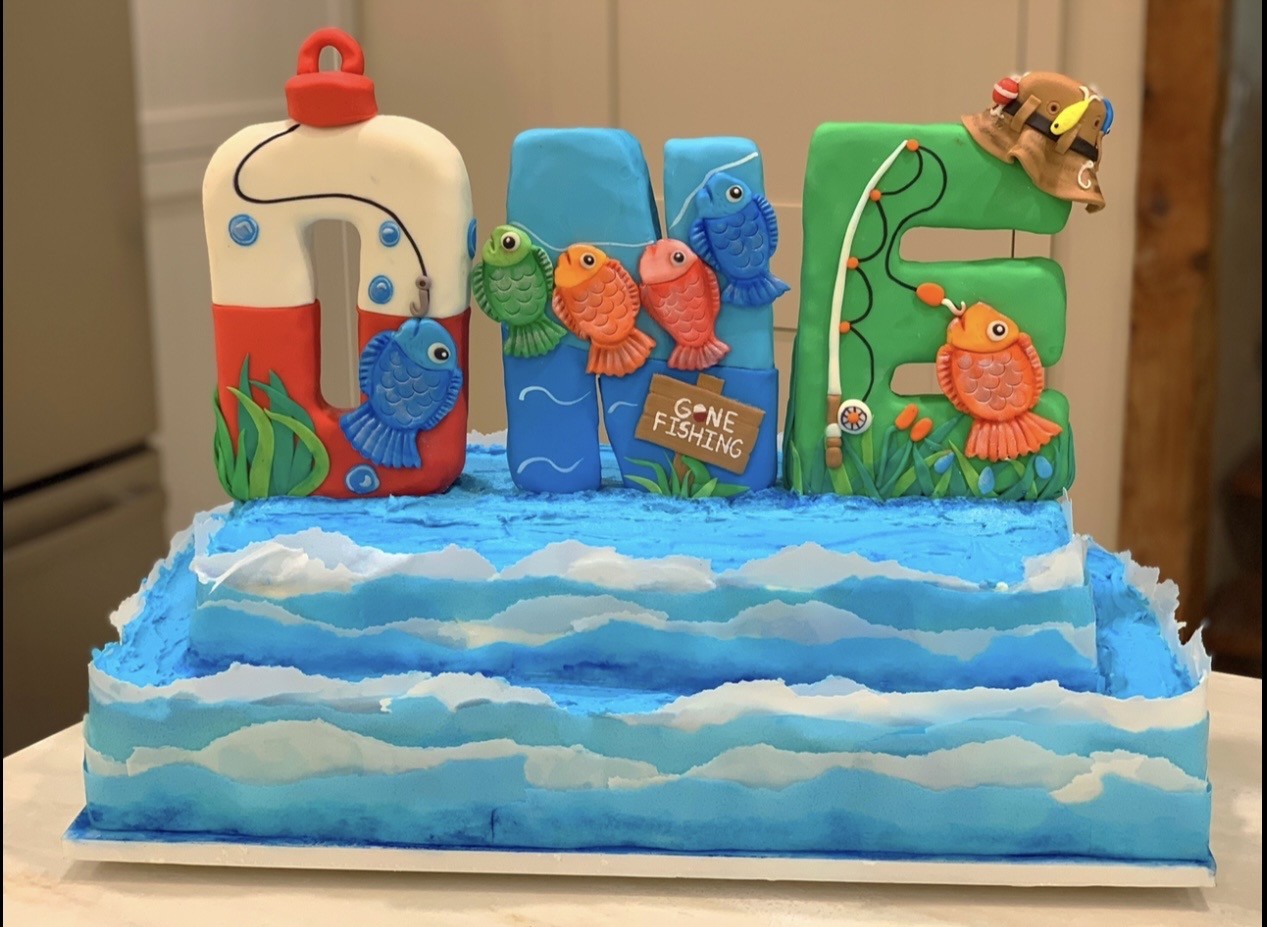 A fishing-themed birthday cake with the name "JOE" spelled out in large sculpted fondant letters each uniquely decorated — the "J" as a red-and-white buoy, the "O" with colorful fish and a "GONE FISHING" sign, the "E" in green with a fishing rod and lily pads — all emerging from a blue and white frosted ocean wave base.