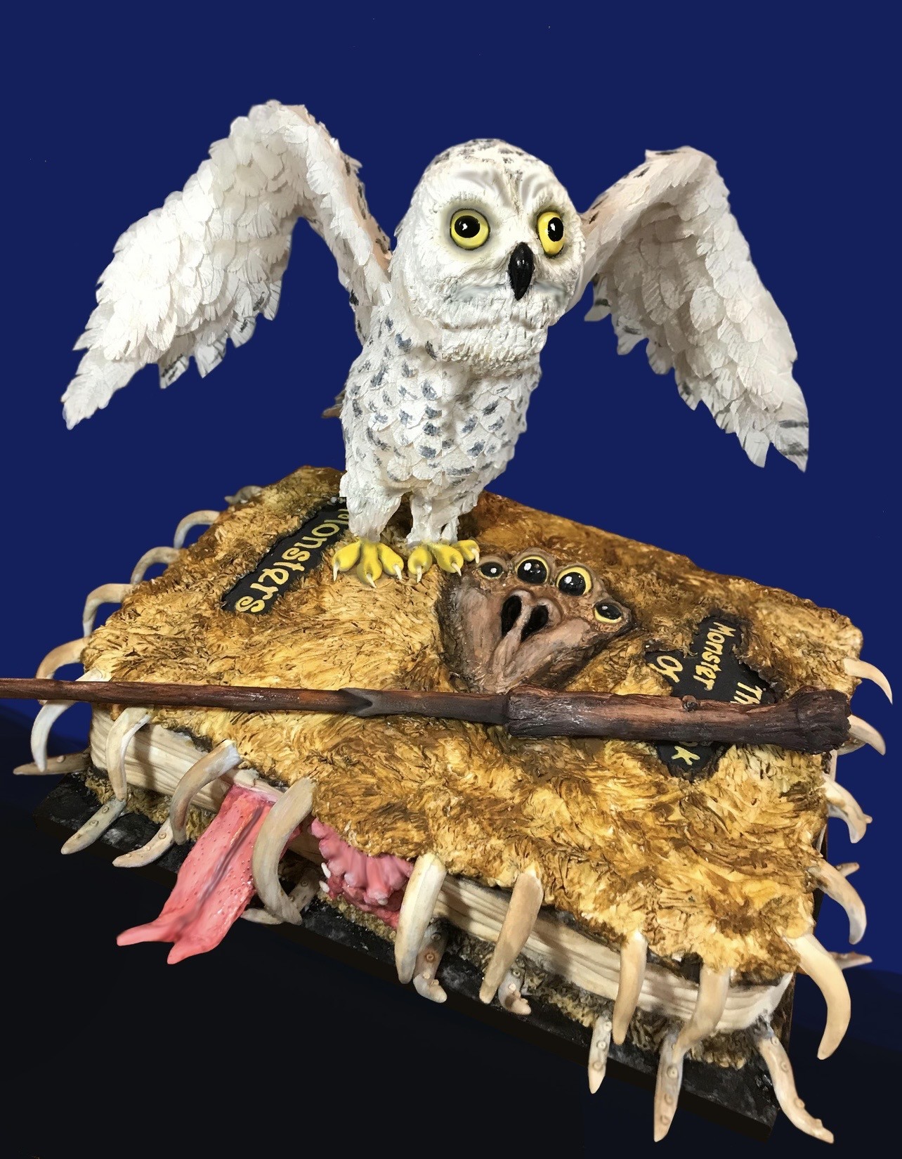 A Harry Potter-themed cake featuring a sculpted white owl resembling Hedwig perched atop a monstrous animated book with a gaping mouth, wooden wand, sharp bone-like teeth, and a long pink tongue, set against a deep blue background.