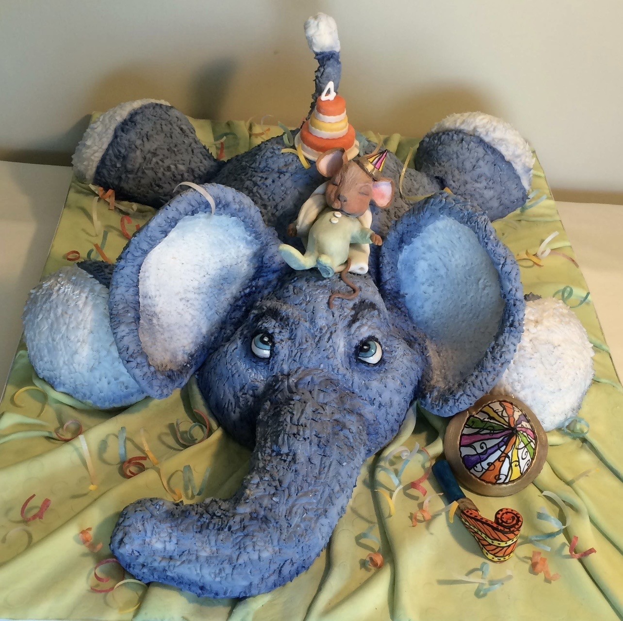 A three-dimensional cake sculpted as Dumbo the elephant in textured blue fondant with soulful blue eyes, a tiny Timothy Q. Mouse figurine wearing a party hat on his head, a striped party hat with the number "4" suggesting a fourth birthday, scattered confetti and ribbon curls, a stained-glass-style circus tent decoration, and a horn-shaped party favor.