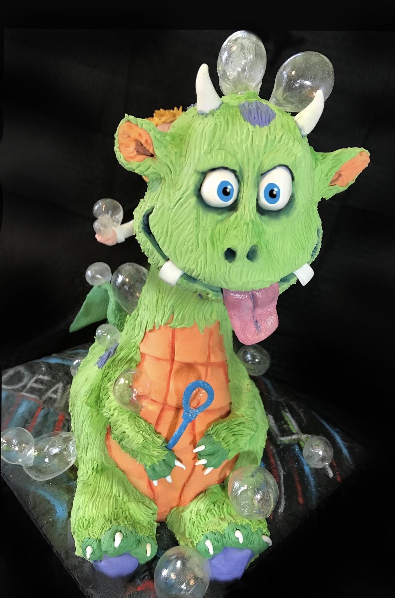 A sculpted cake of a cartoonish green dragon with textured fondant skin, orange segmented belly, purple accents on feet and forehead, large expressive blue eyes, and a playful tongue, holding a small blue bubble wand with translucent bubble-like spheres scattered around, perched on a dark chalkboard-style base.