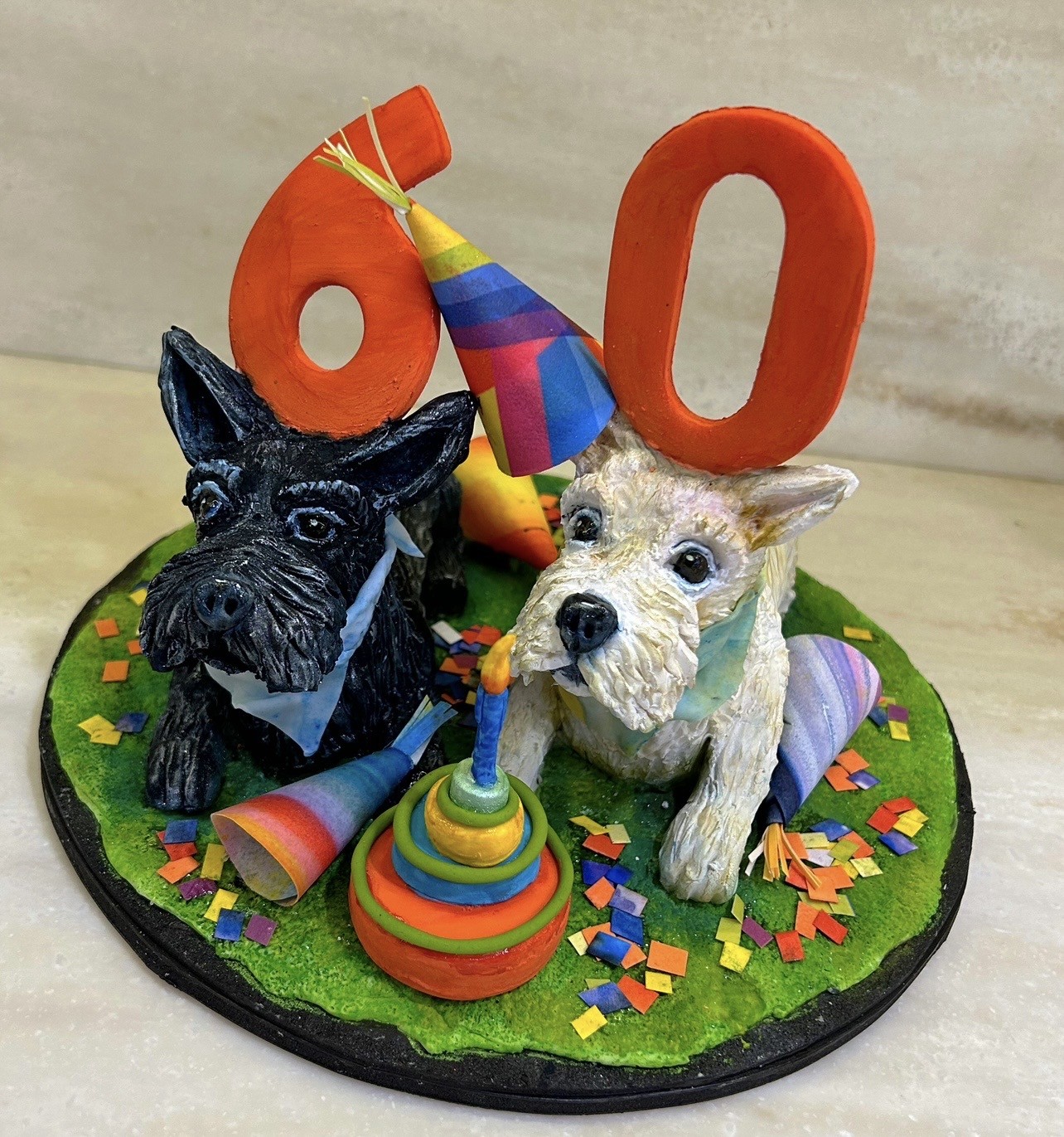 A 60th birthday cake topper featuring two sculpted dogs — a Scottish Terrier in black with light blue bow tie and a West Highland White Terrier in white with rainbow-striped party hat — positioned on green grass-like base with multicolored confetti, between them a small tiered birthday cake with single lit candle.