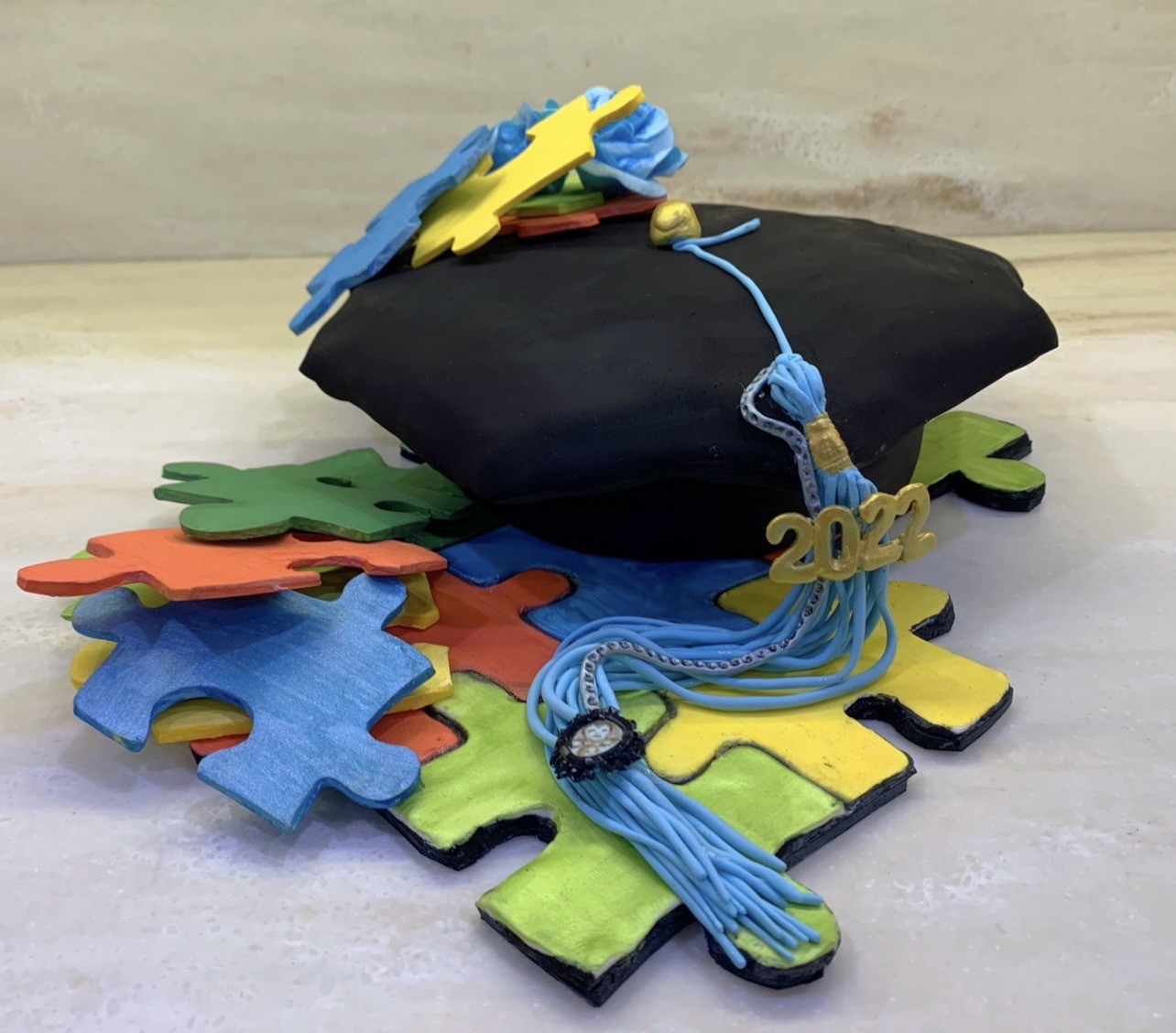 A sculpted cake of a graduation cap in black mortarboard with light blue tassel cascading over colorful puzzle pieces, gold-colored agate and decorative bead on tassel, golden "2022" numeral on the side, abstract shapes in blue and yellow on top of the cap.