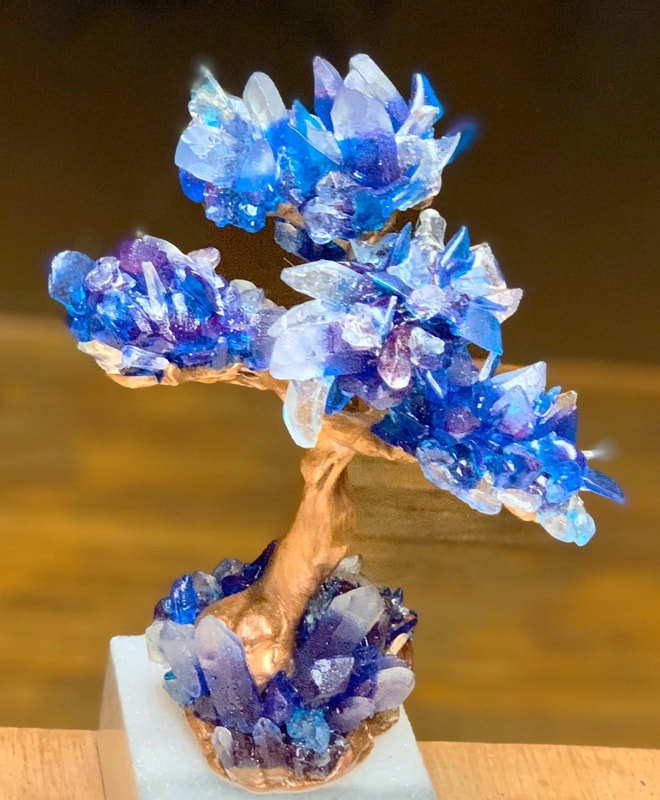 A handcrafted sugar bonsai-style tree sculpture features a twisted copper-colored wire trunk and branches adorned with clusters of blue and purple sugar quartz crystals, mounted on a white rectangular base.