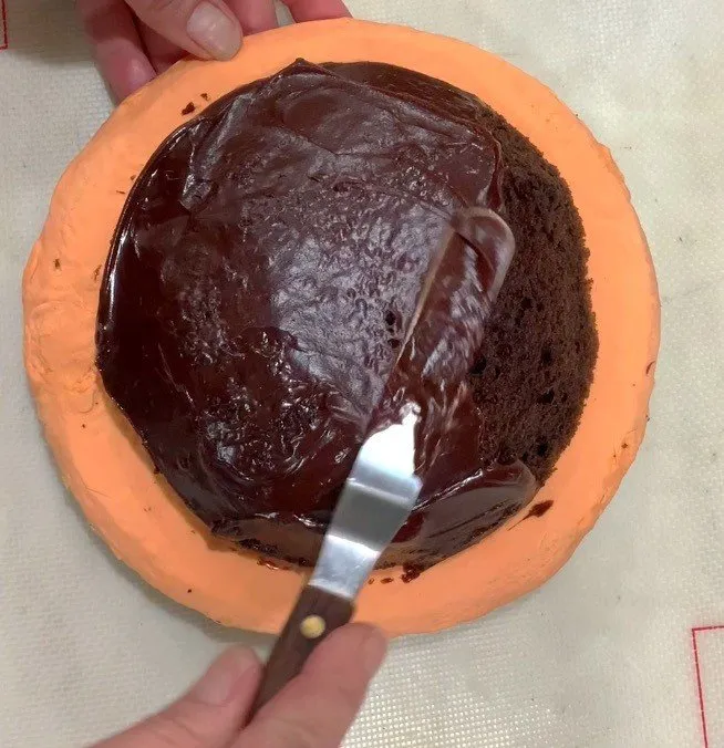 A hand spreads chocolate ganache over a carved cake layer with an offset spatula.