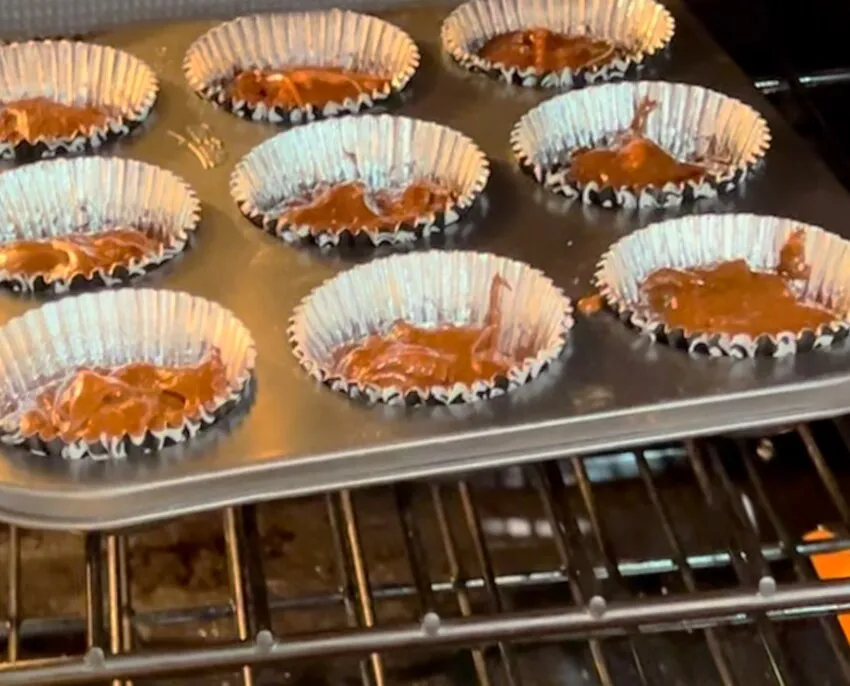 A muffin tin of chocolate cupcakes baking on a wire rack, still in their paper liners