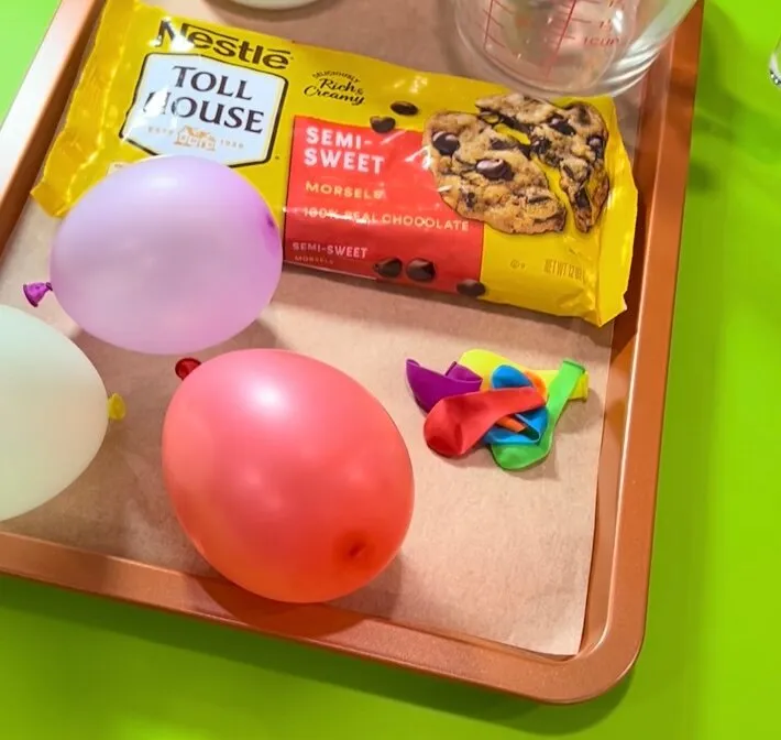 Ingredients and supplies for making chocolate balloon cups laid out on a baking tray atop a bright green surface