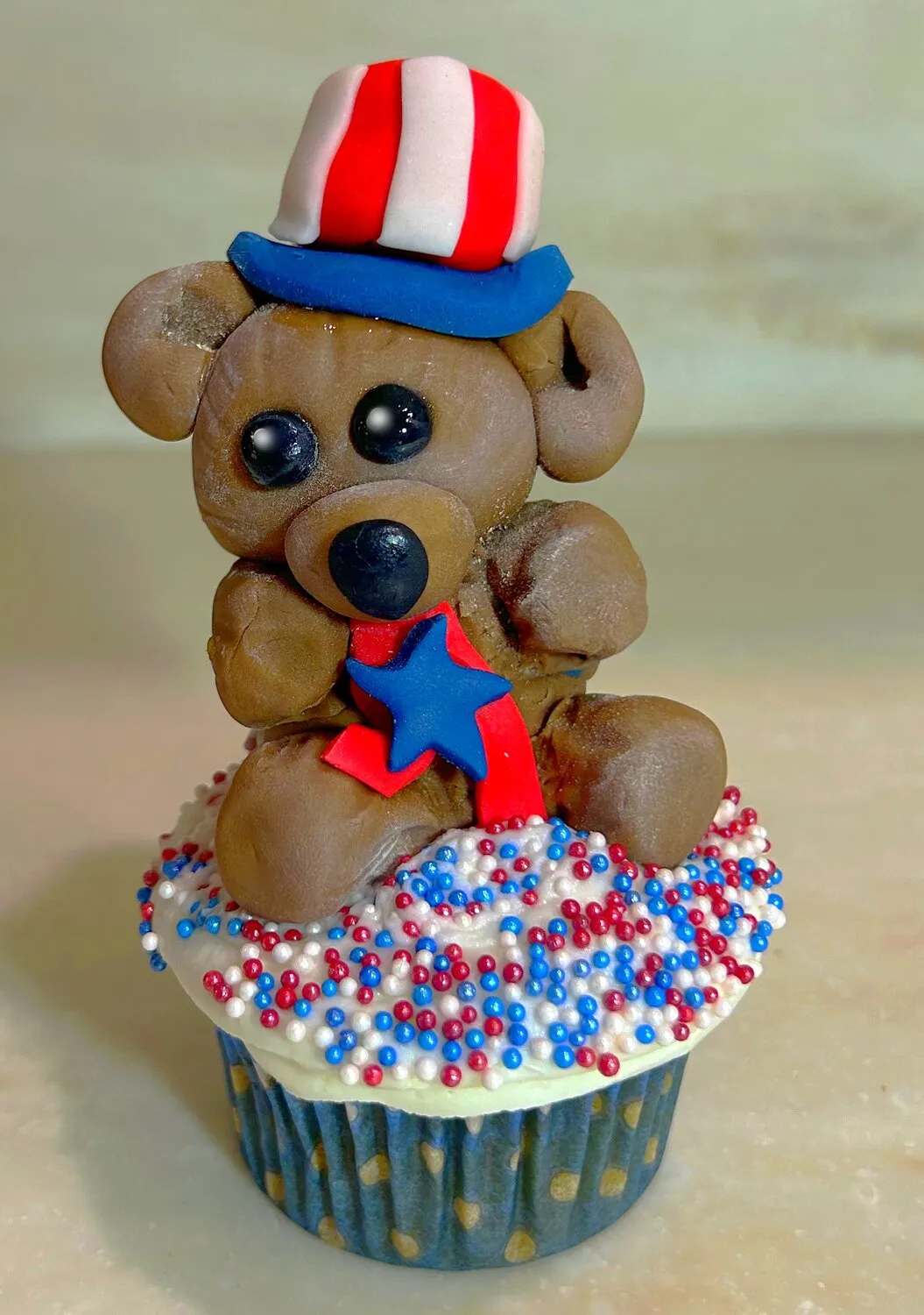 A red, white and blue teddy bear made from fondant sitting on a cupcake on a wooden surface