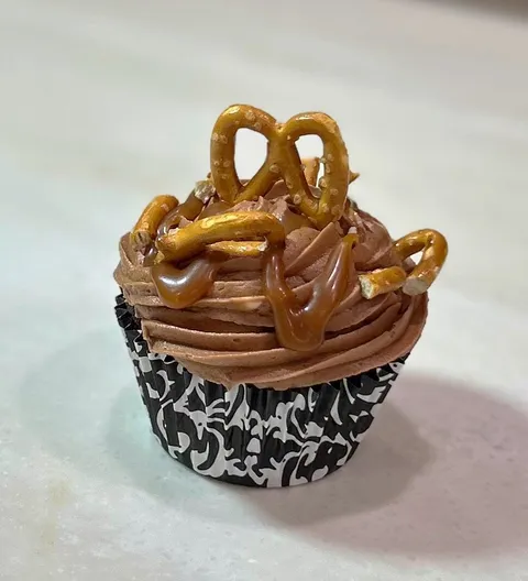 Chocolate cupcake with chocolate buttercream frosting, caramel drizzle, and pretzel pieces as garnish, sitting in a black and white patterned cupcake liner on a light-colored surface