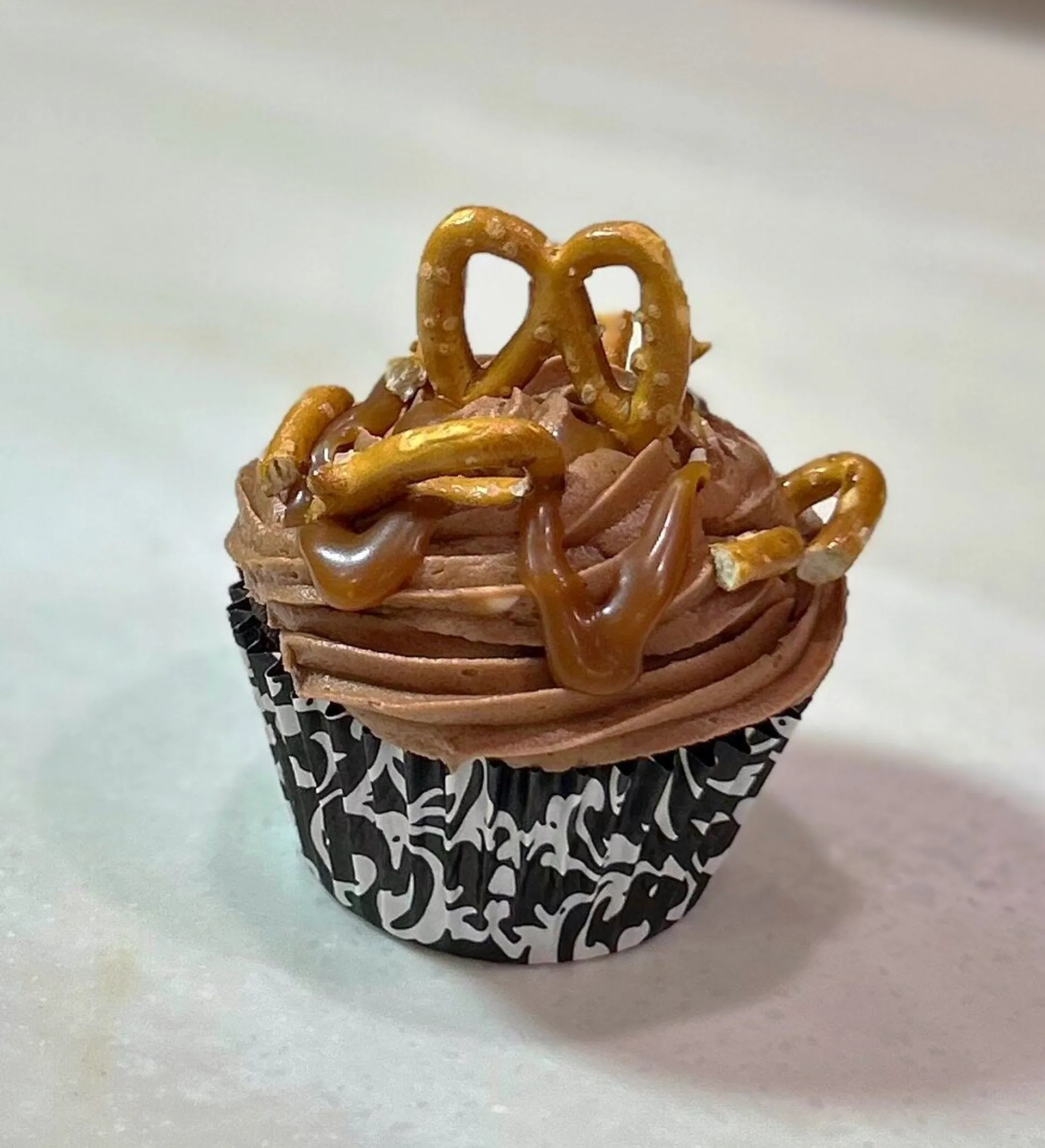Chocolate cupcake with chocolate buttercream frosting, caramel drizzle, and pretzel pieces as garnish, sitting in a black and white patterned cupcake liner on a light-colored surface