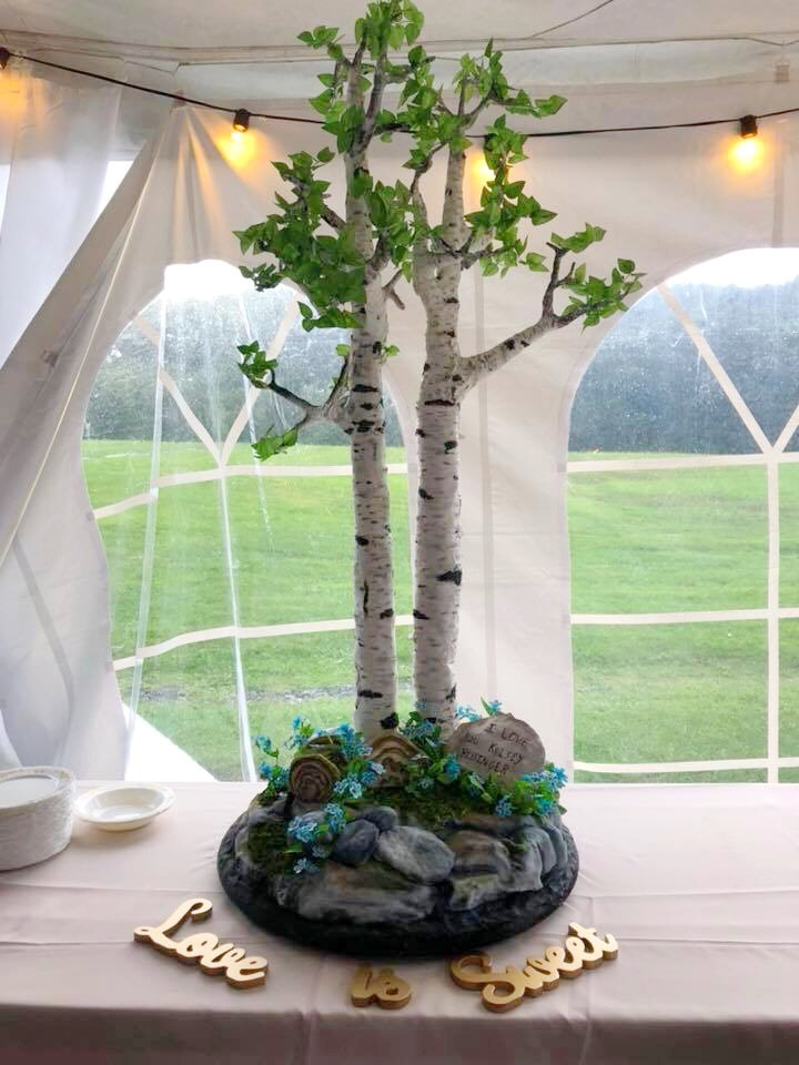A tall, sculpted birch tree centerpiece with green leaves and a rocky base decorated with blue flowers sits on a table inside a white tent, accompanied by wooden letters spelling "Love is Sweet."