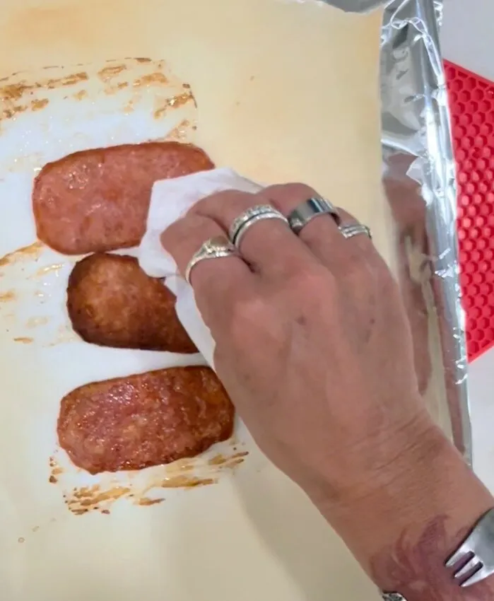 Hand blotting fried meat patties with a paper towel on a baking sheet.