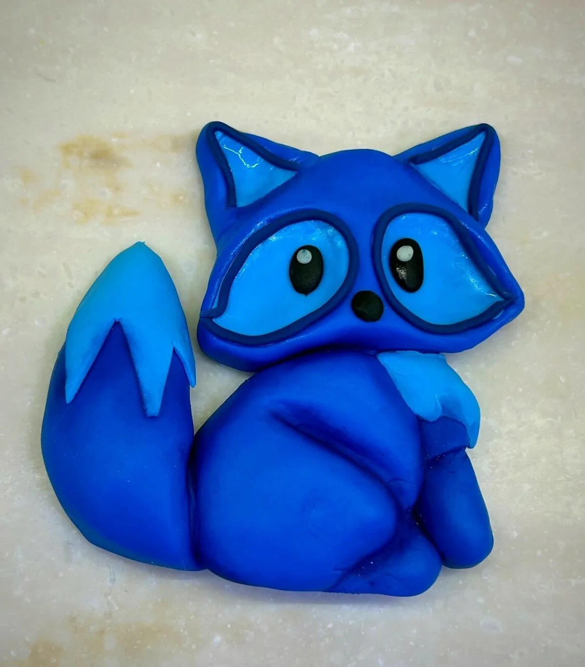 A blue fondant fox sugar piece with large eyes and a fluffy tail on a light surface