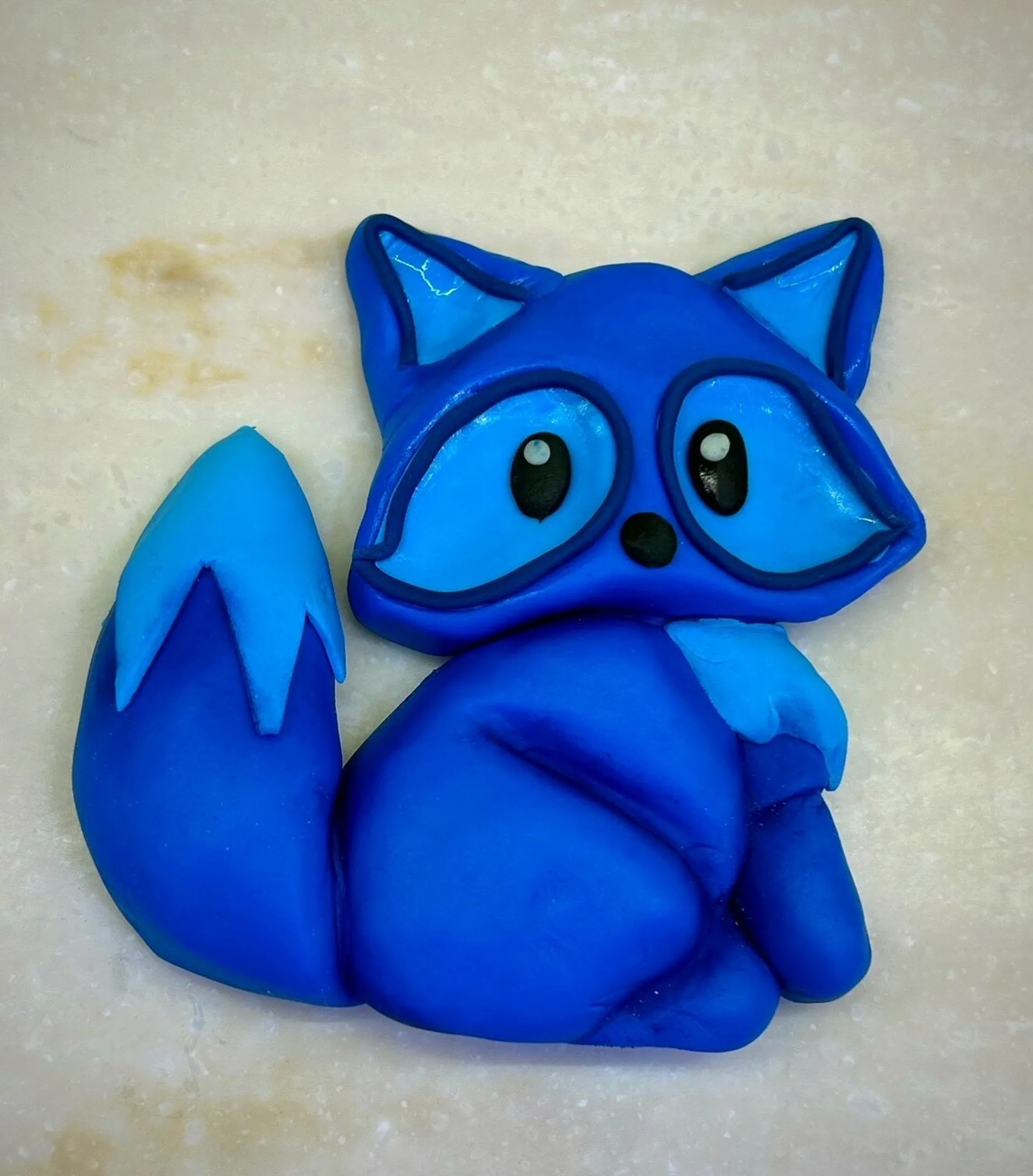 A blue fondant fox sugar piece with large eyes and a fluffy tail on a light surface