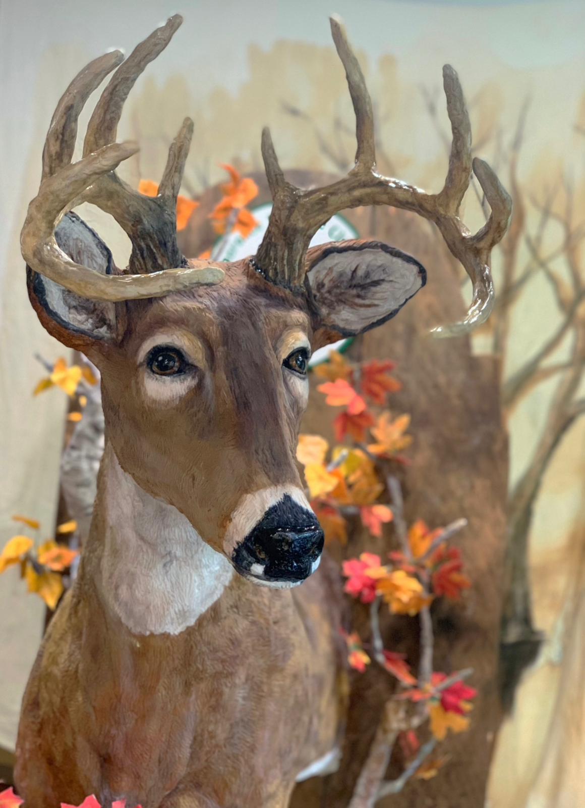 A realistic sculpted deer cake with large antlers and a black nose, shown in a close-up view against a painted autumn forest backdrop featuring orange and yellow leaves.