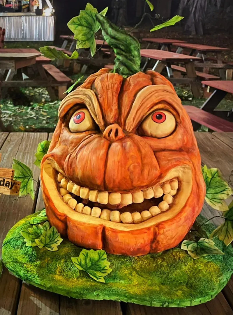 A grotesque, hyper-realistic jack-o'-lantern with bulging red eyes, a pig-like snout, and a wide, toothy grin sits on a mossy green base surrounded by ivy leaves, with a small wooden sign reading "day" visible to the left.