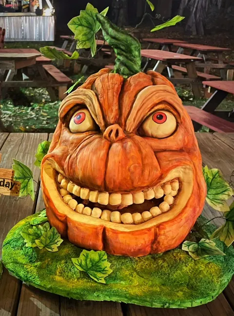 A grotesque, hyper-realistic jack-o'-lantern with bulging red eyes, a pig-like snout, and a wide, toothy grin sits on a mossy green base surrounded by ivy leaves, with a small wooden sign reading "day" visible to the left.