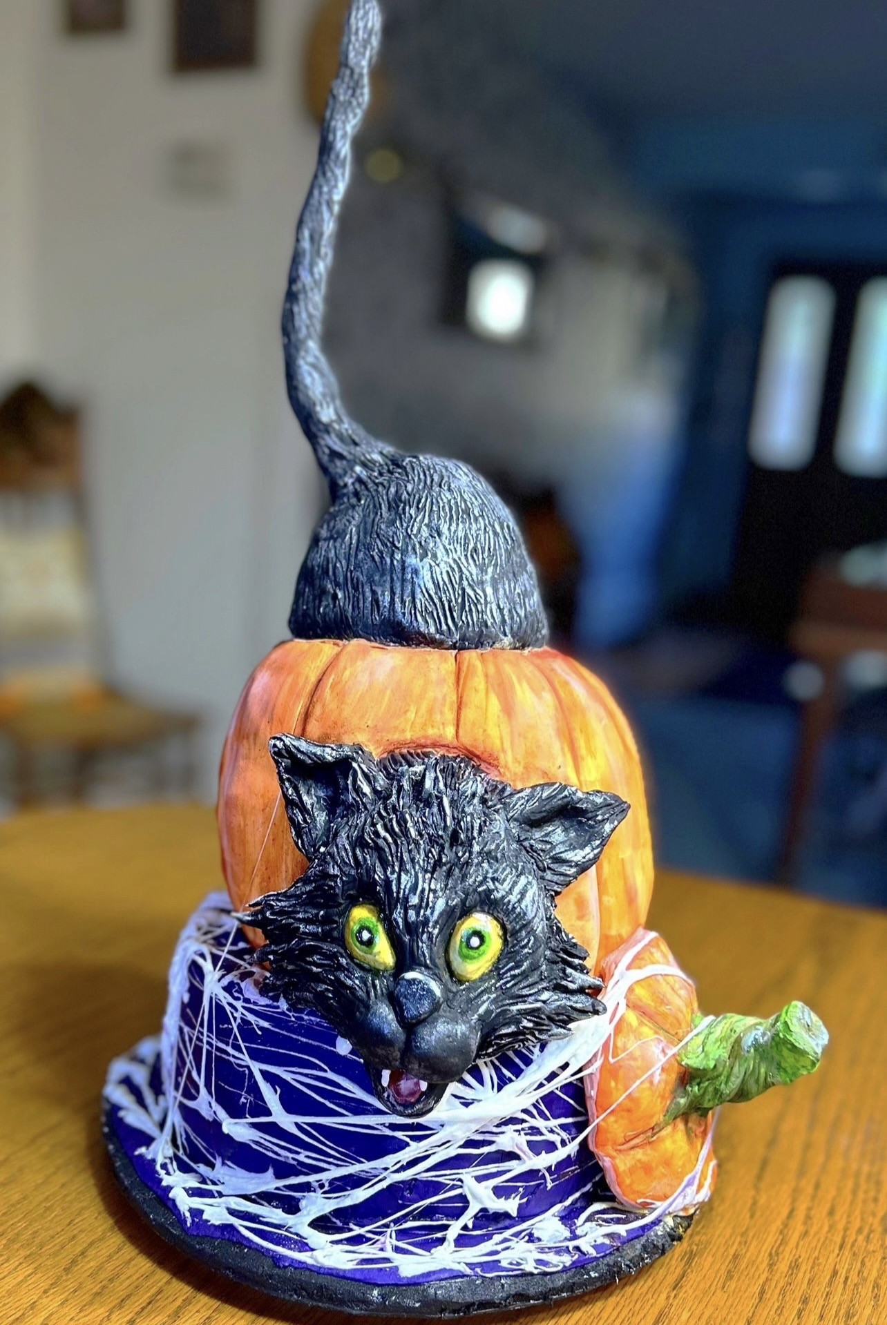 A whimsical Halloween sculpted cake featuring a glossy black cat with vivid green eyes and open mouth leaping from behind a bright orange pumpkin forming the upper body, the cat's tail arching dramatically curling above the pumpkin, set on a base resembling a witch's hat in deep purple with white spiderweb patterns.