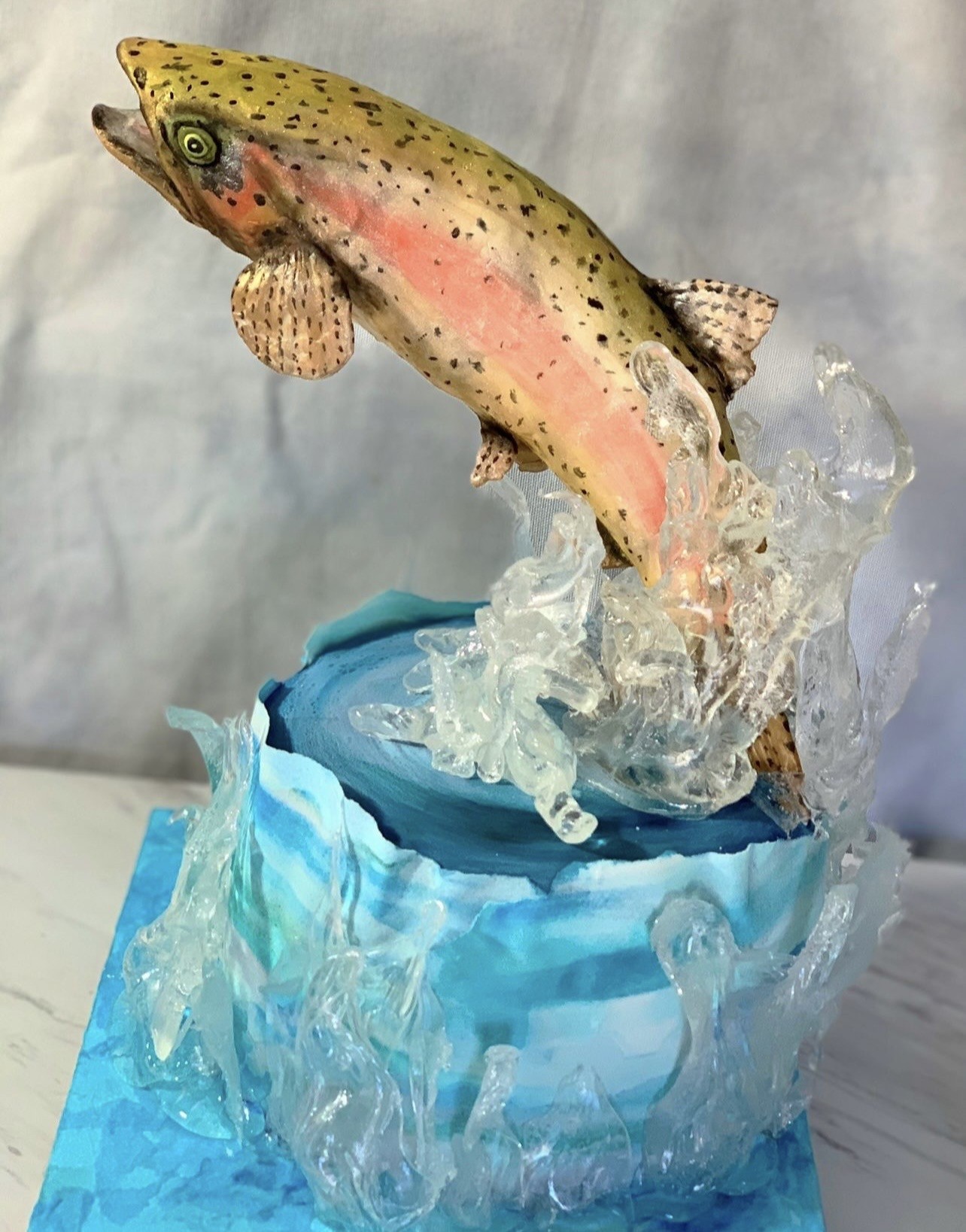 A sculpted cake of a leaping trout captured mid-air breaking through stylized water, the fish rendered with golden-olive back speckled with black spots, vibrant pink lateral stripe, and translucent fins, emerging from a dynamic splash of clear resin-like material mimicking frothy water atop a cylindrical base painted in swirling blue and turquoise.