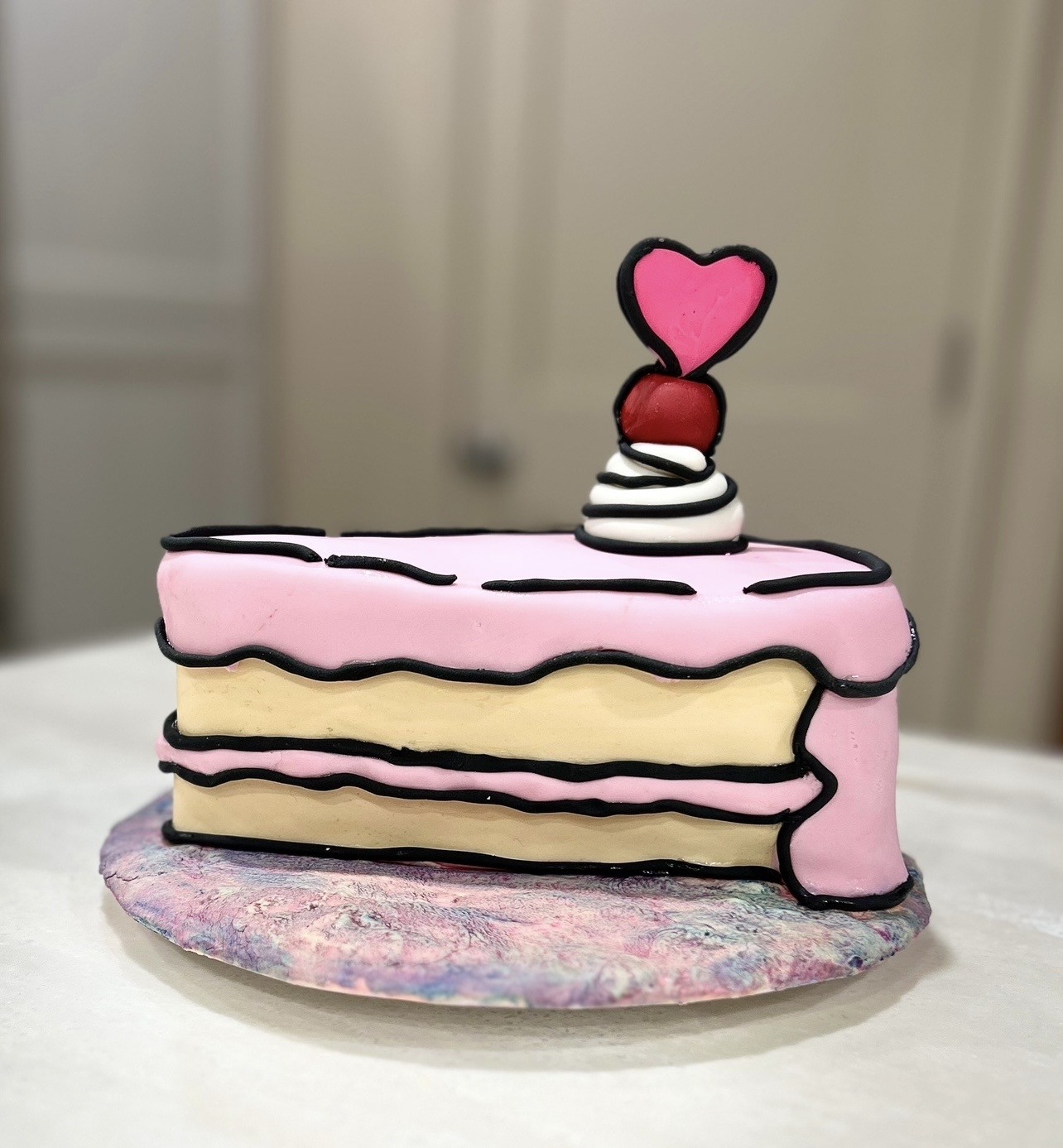 A whimsical cake designed as a slice of layered dessert in cartoonish comic-book style, with alternating horizontal layers of pale yellow sponge and soft pink frosting outlined in bold black lines, topped with white whipped cream, a glossy red cherry, and a pink heart-shaped topper.