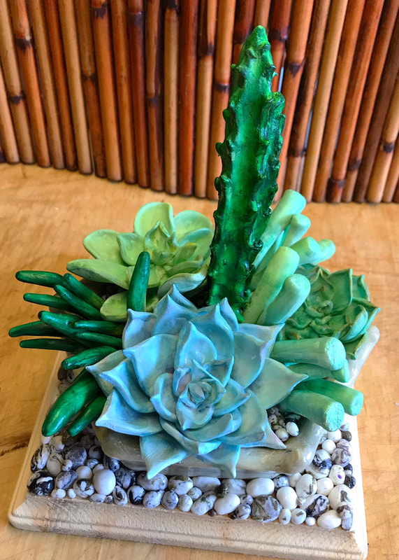 A handcrafted clay sculpture of a succulent garden, featuring various green and blue-green cacti and rosettes planted in a bed of small white and grey pebbles within a square planter.