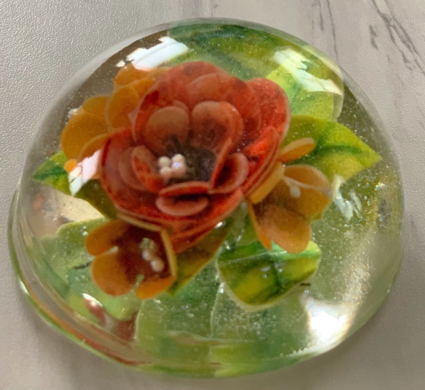 A clear sugar glass paperweight encasing a detailed orange and red sugar flower surrounded by green leaves.