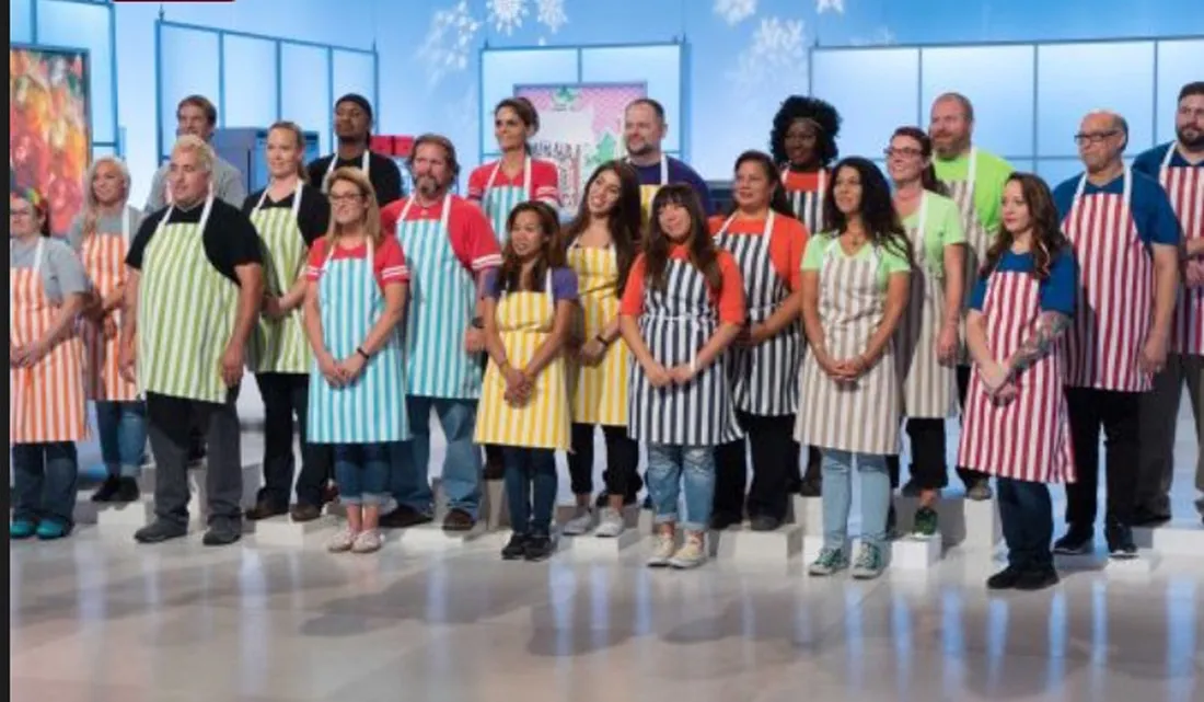 Contestants in striped aprons on tiered platforms on the set of Cake Wars Christmas.
