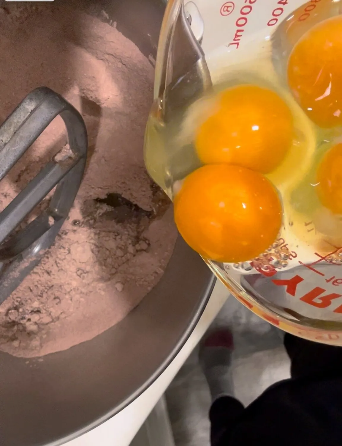 A stand mixer bowl with cake mix and pudding powder, eggs being poured in from a glass measuring cup