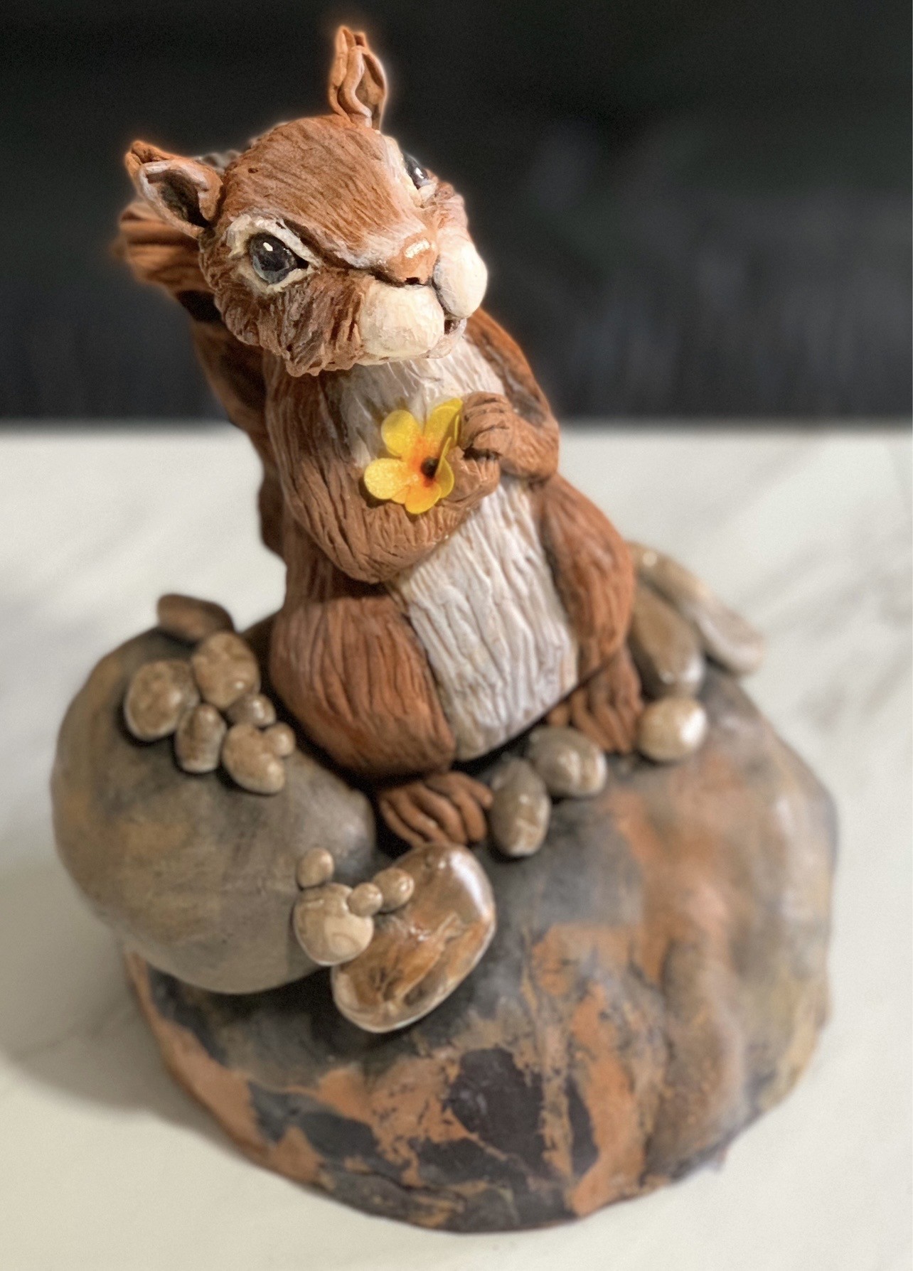A sculpted cake figurine of a squirrel seated upright on a textured organic base resembling a weathered rock, with finely detailed chestnut brown fur, creamy white underbelly, glossy black eyes, holding a small bright yellow flower with an orange center.